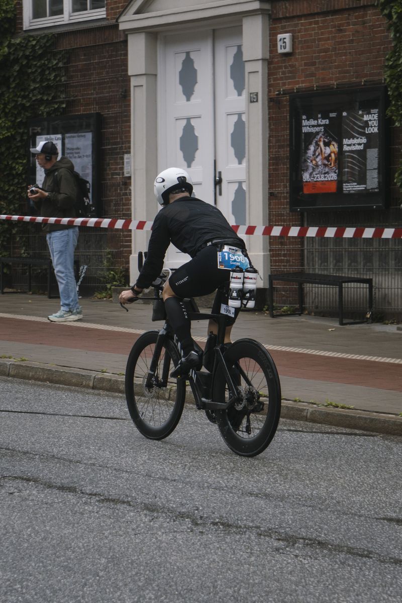 Ein Triathlet fährt von hinten gesehen durch eine Wohnstraße, ein Passant schaut vom Gehweg aus zu