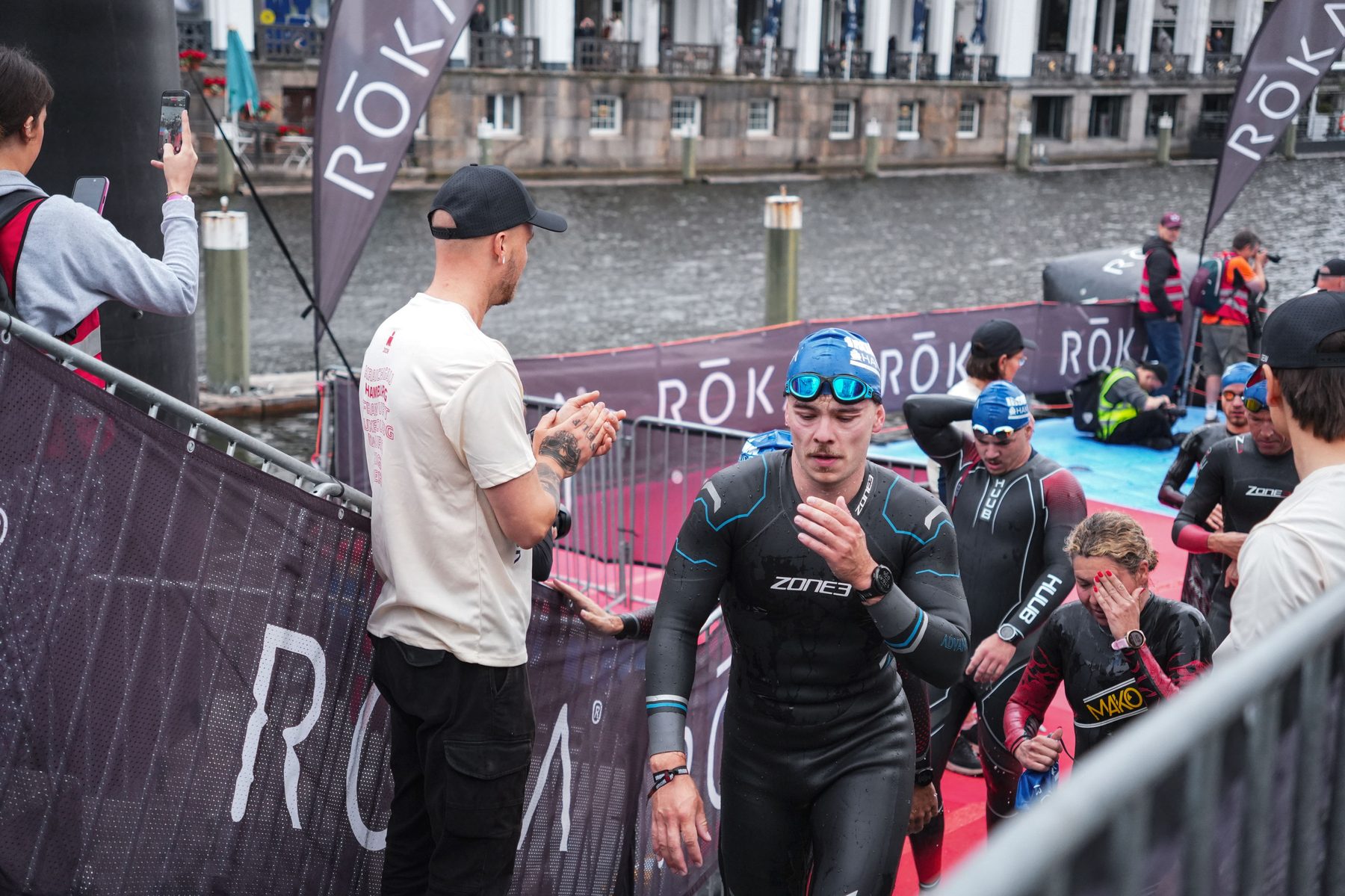 Triathleten im Neoprenanzug auf dem Weg zum Schwimmstart an der Alster, ein Zuschauer klatscht Beifall