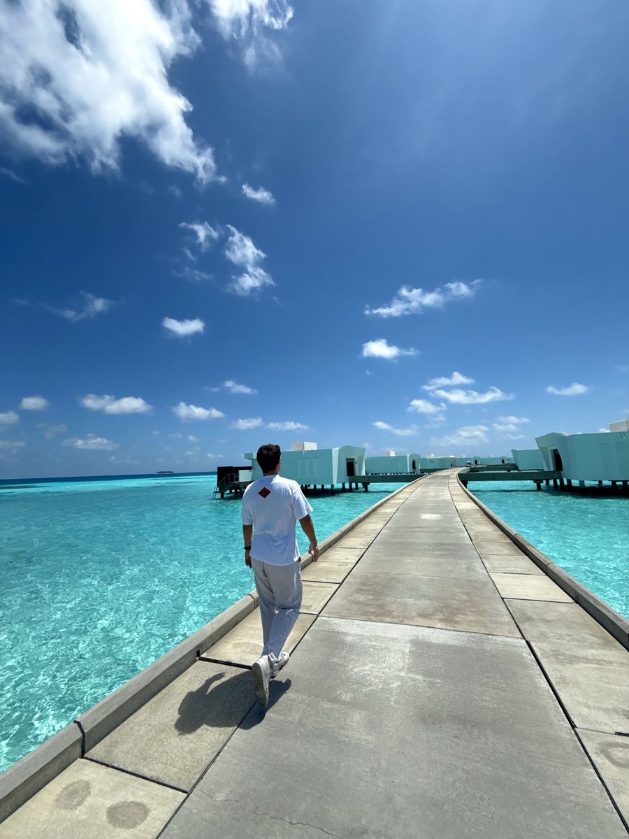 Ein Mann geht über einen langen Steg, der durch türkisfarbenes Wasser zu Wasserbungalows führt