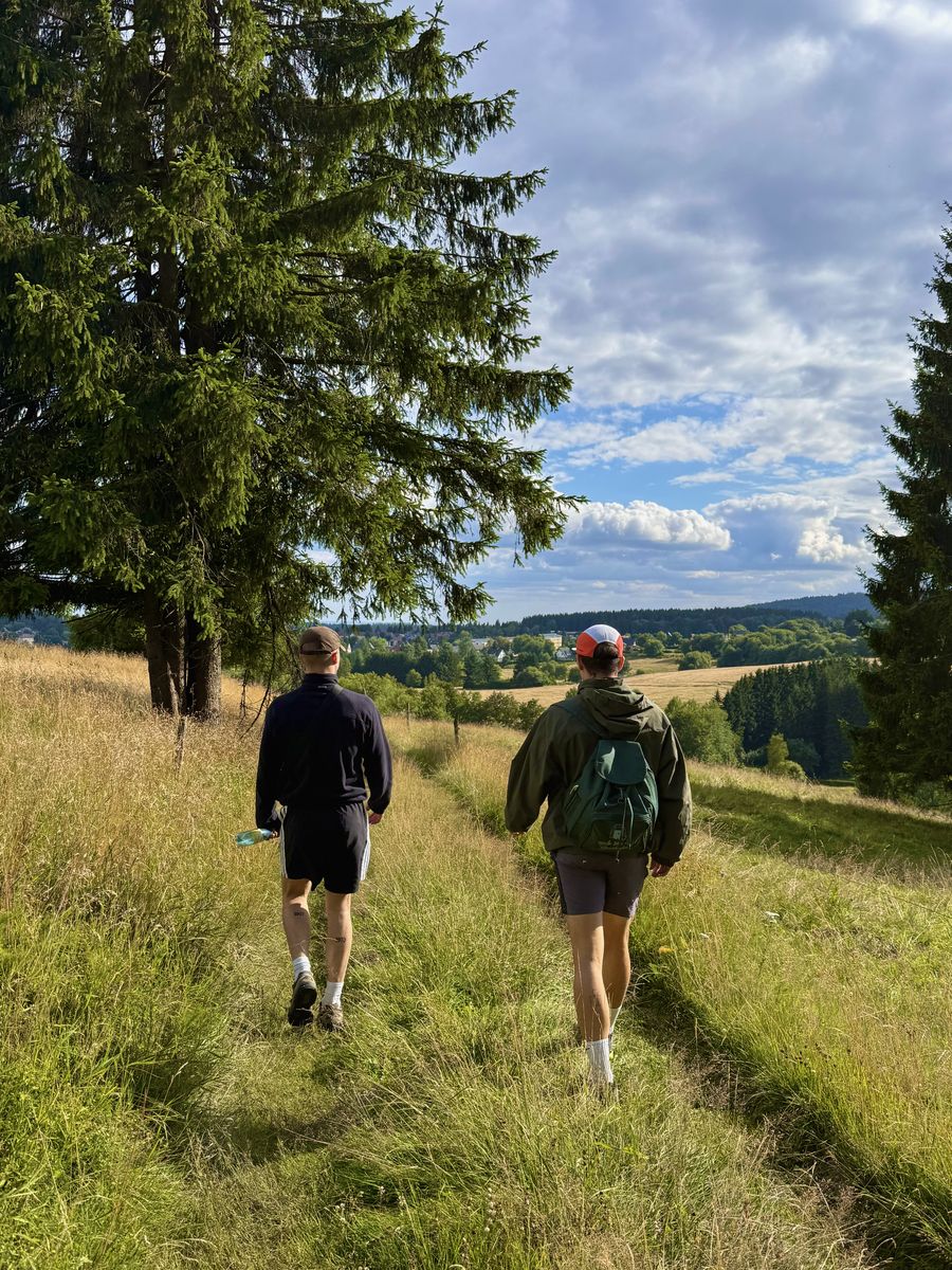 Zwei Wanderer gehen nebeneinander einen Wiesenpfad hinauf, dahinter bewaldete Hügel unter Wolken