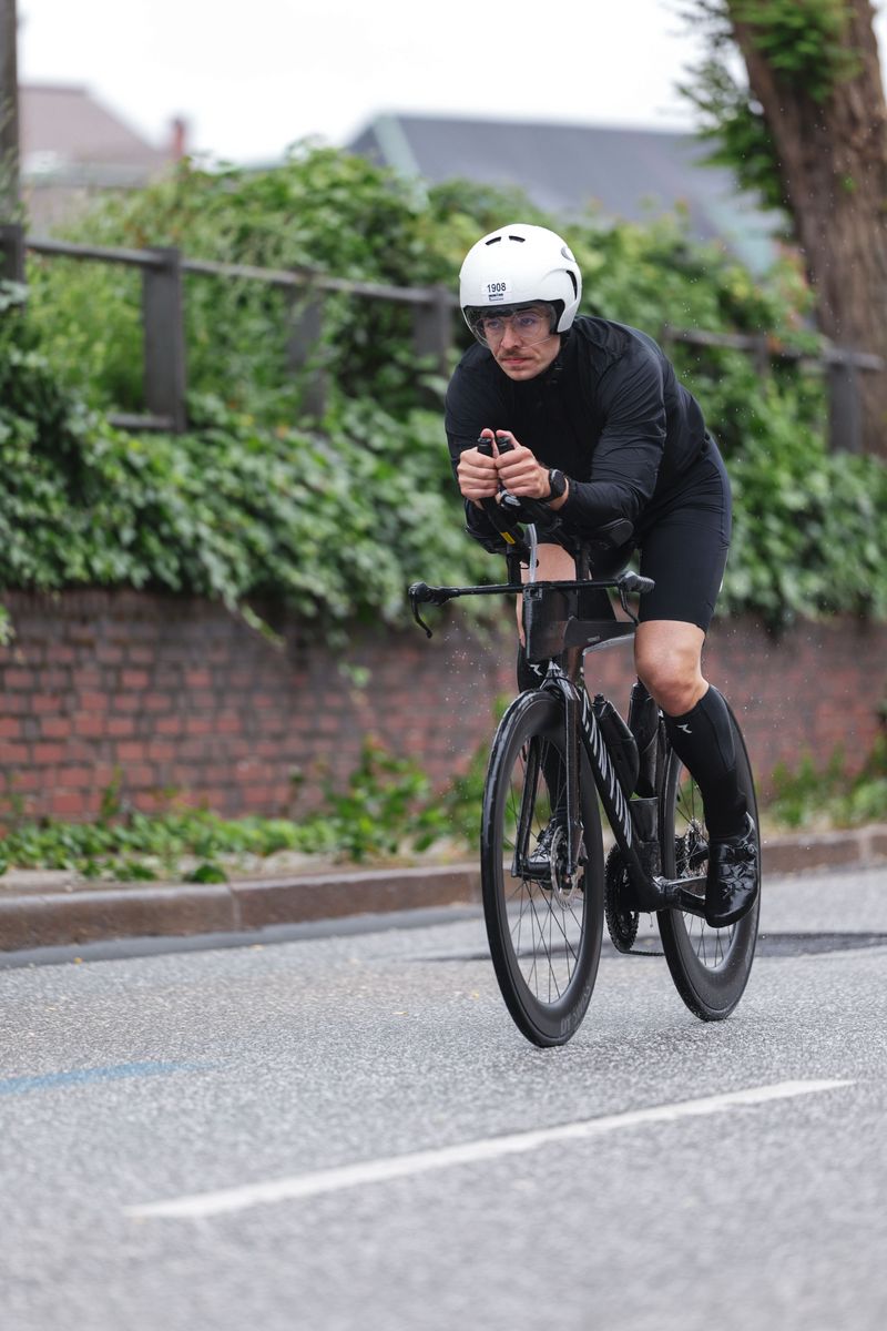 Lucas liegt im Aero-Lenker auf dem Rad, weißer Zeitfahrhelm, dahinter eine Backsteinmauer